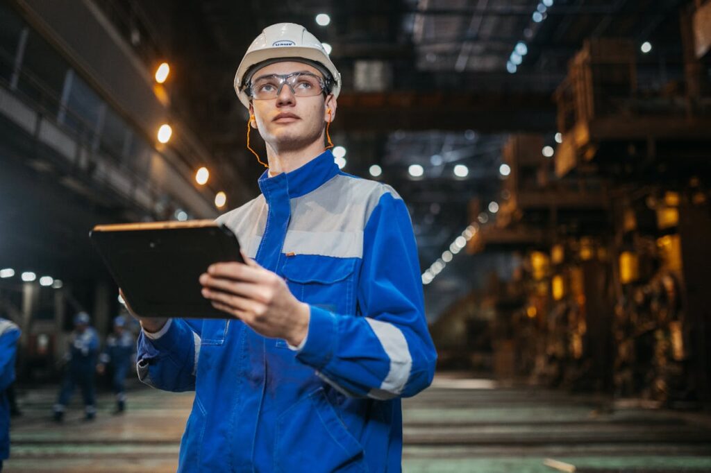 pexels-photo-32845694 Engineer in safety gear uses a tablet in a large industrial factory setting, focused and diligent.